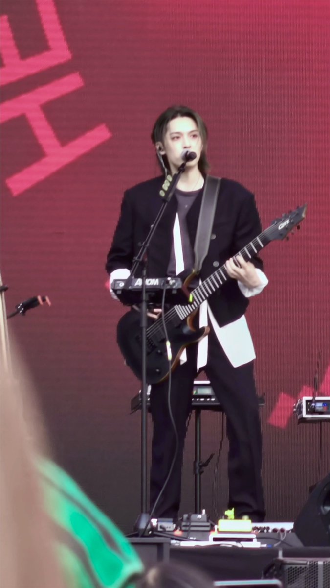 A musician in a black suit performing on stage, playing an electric guitar and singing into a microphone against a pink LED background.