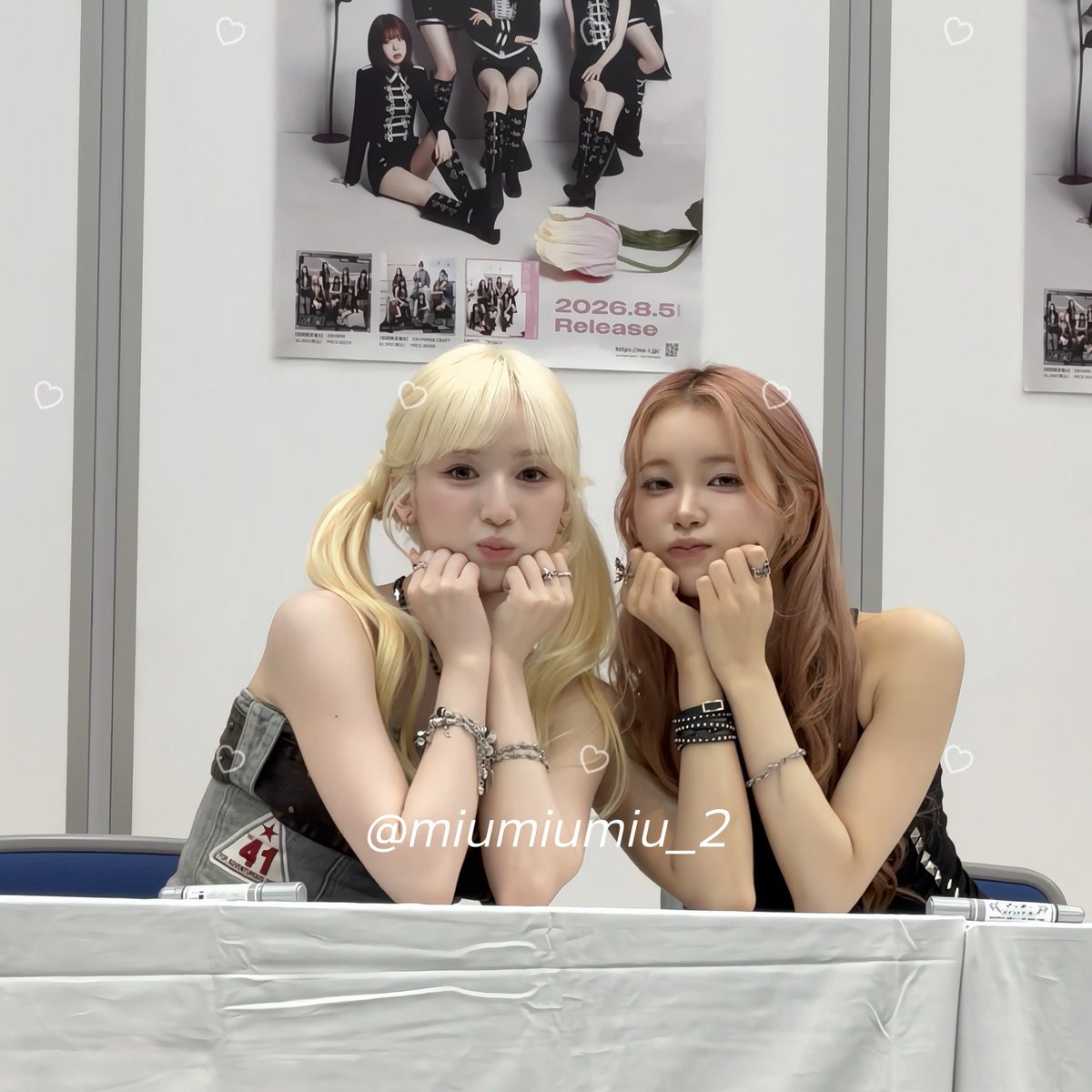Two young women with blonde and pink hair pose closely together with their hands cupped under their chins, smiling sweetly at the camera. Behind them is a promotional poster dated "2026.8.5 Release," and a watermark reads "@miumiumiu_2," capturing a sweet moment from the ME:I Kansai Offline All-Member Signing Event featuring Miu and Suzu.