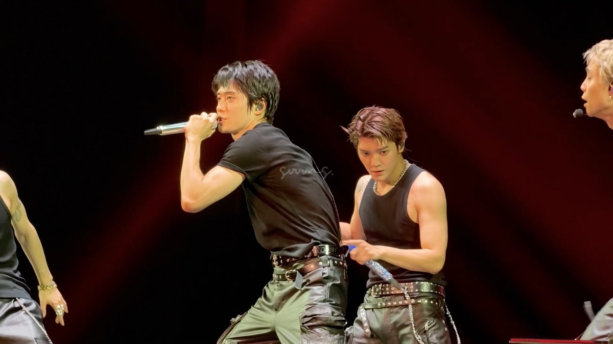 A concert photograph showing performers on stage, including a singer in a black t-shirt with a microphone and backup performers, with a watermark 'SunnyS.' visible.