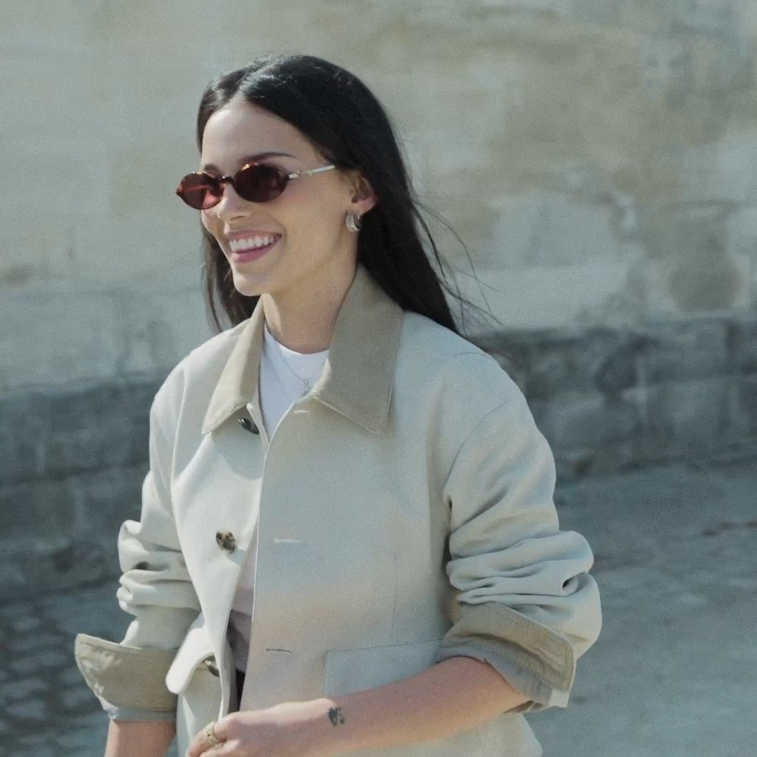 A woman with long dark hair, wearing tortoiseshell sunglasses and an oversized beige trench coat with contrasting tan collar and cuffs, smiles brightly outdoors against a stone wall backdrop, capturing the elegant, sun-drenched aesthetic of the Dior Summer 2027 Women's show.