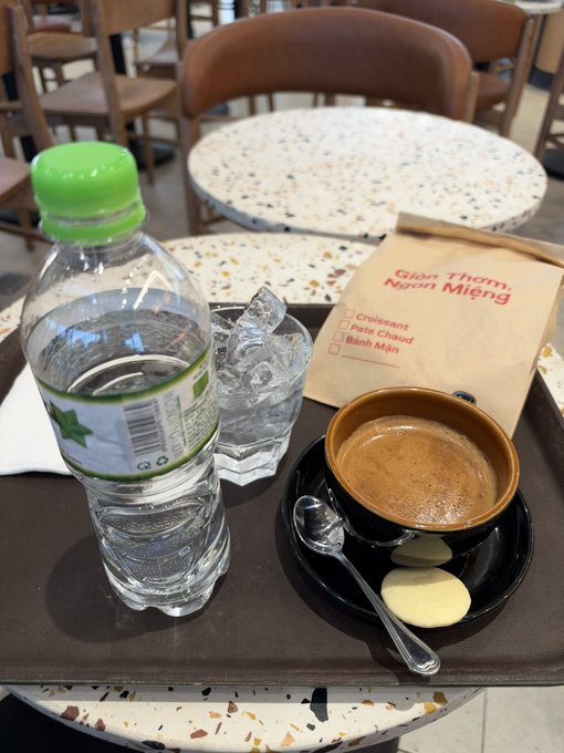 A bottle of water, a glass of ice water, a cup of coffee on a saucer with cookies, and a paper bag labeled "Gim Thom Nhạn Mieng" with options for Croissant, Pate Chaud, and Banh Mi sit on a tray at a cafe.
