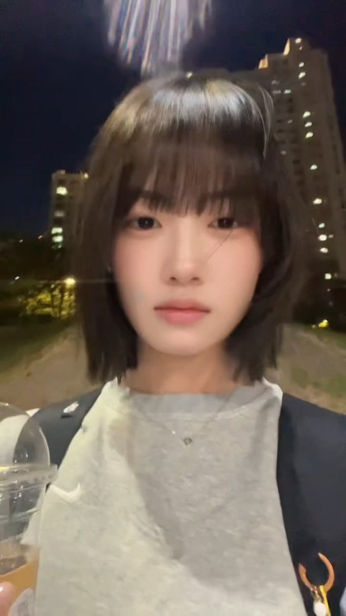A nighttime selfie of a young woman with short brown hair and bangs wearing a grey t-shirt, taken outdoors near apartment buildings.