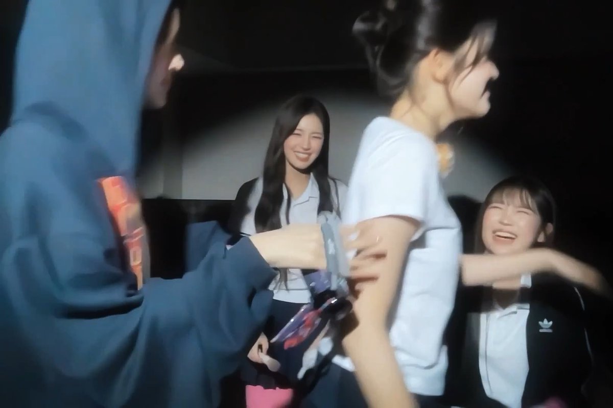 A blurry action photo capturing people laughing and moving, with a woman in a white shirt smiling in the background while others in the foreground appear to be in motion.