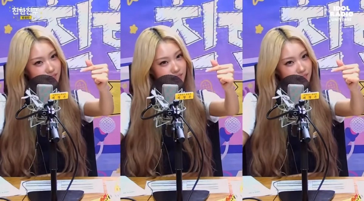 A blonde young woman sits at a microphone in a radio studio with 'IDOL RADIO UNIVERSE' visible, wearing a navy and white outfit and making finger heart gestures.
