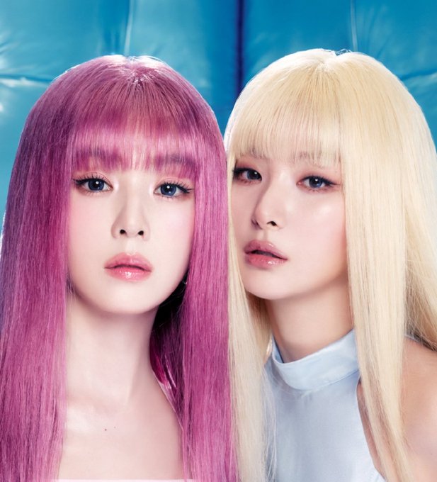 Two young women pose closely together, one with long pink hair and the other with platinum blonde hair, against a bright blue backdrop.