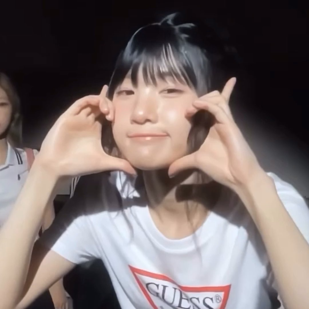 A young woman in a white Guess t-shirt with bangs smiles and holds her hands up to form a heart shape around her face, with another person partially visible in the background.