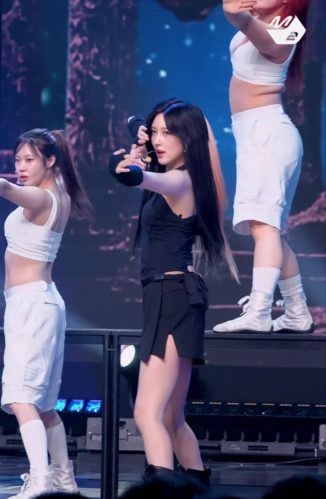 A K-pop performance featuring a female singer in a black outfit holding a microphone, backed by dancers in white tops and shorts on a stage with blue lighting.