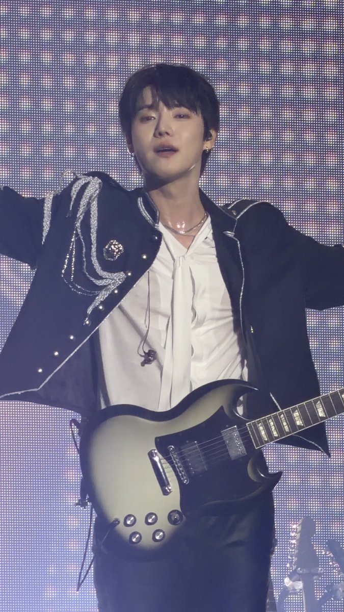 A young male musician performing live, dressed in a black jacket with silver embellishments over a white ruffled shirt, playing a black and cream electric guitar.