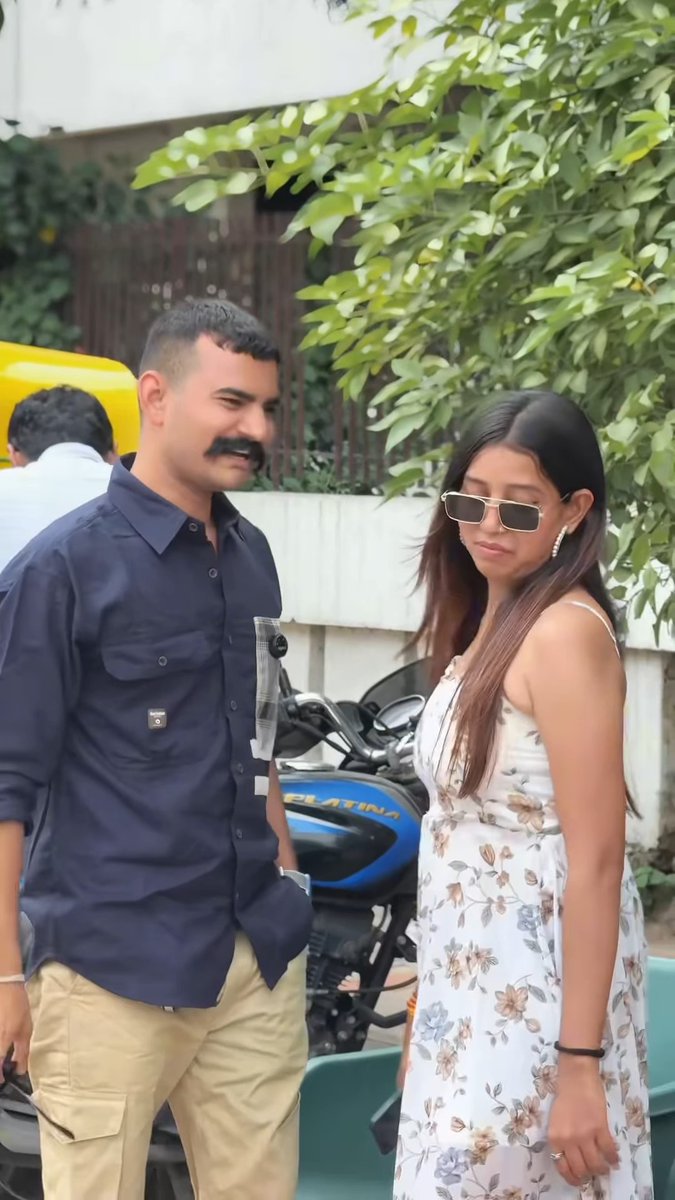 A man with a mustache in a blue shirt smiles at a young woman wearing sunglasses and a white floral dress standing outdoors near a blue 'PLATINA' motorcycle.