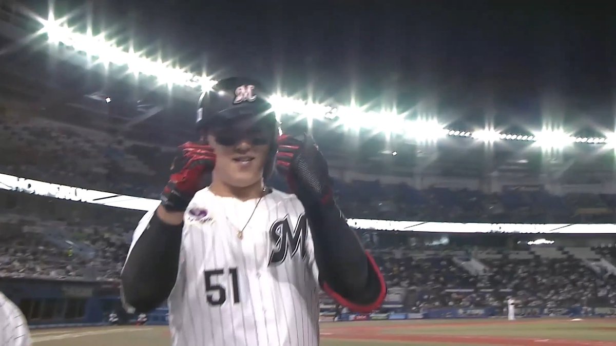 A baseball player wearing a black helmet and jersey number 51 with an 'M' logo adjusts his batting gloves under bright stadium lights, with a crowd visible in the stands behind him.