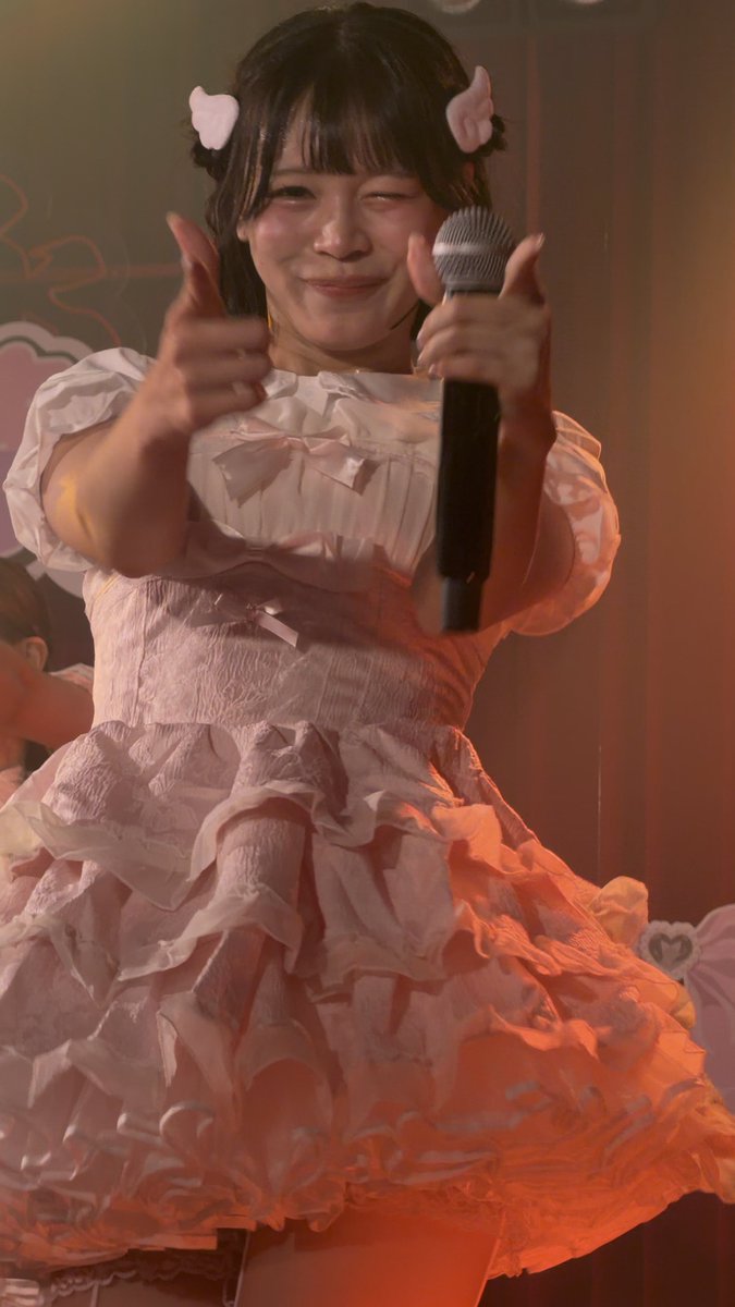 A young woman in a frilly pink dress performs on stage, winking and giving two thumbs up while holding a microphone, with small white wing clips in her hair.