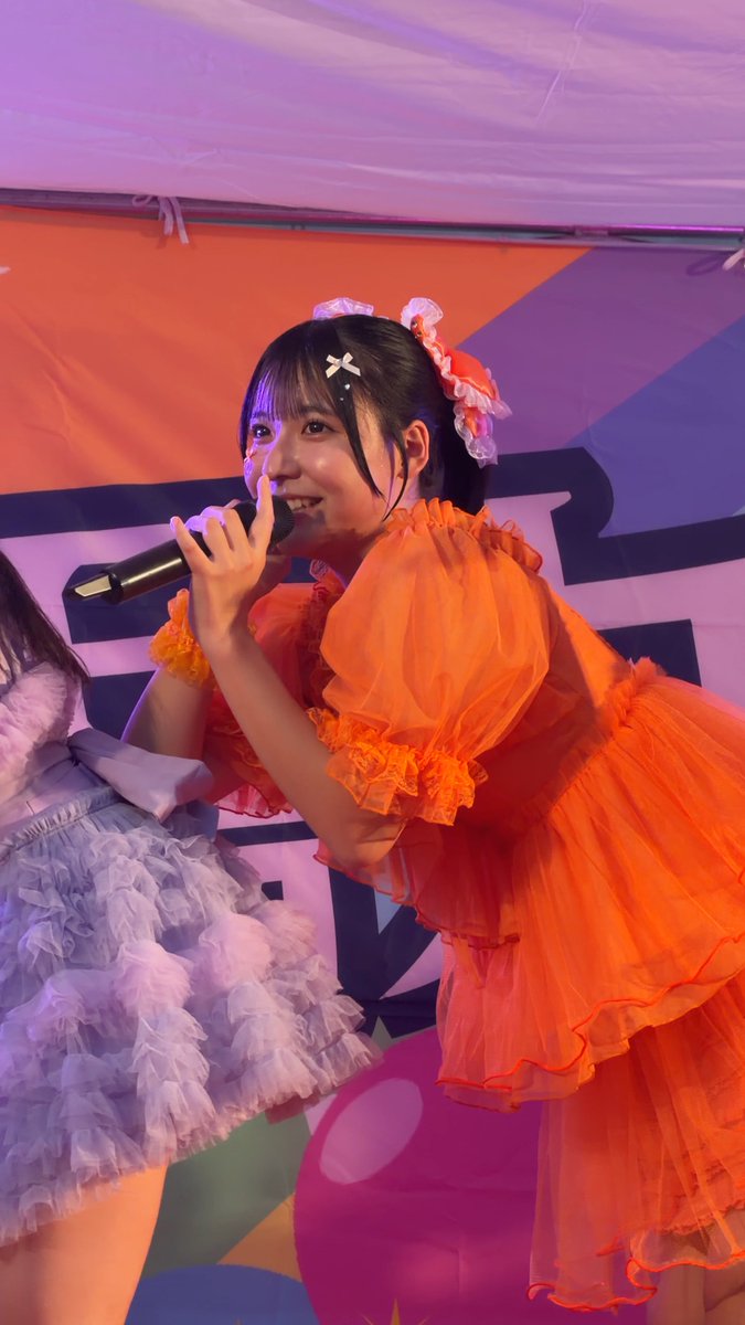 A Japanese idol in a bright orange ruffled dress with hair accessories sings into a microphone on stage.