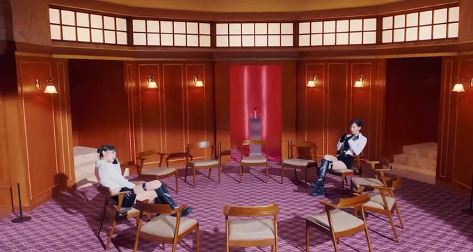 Two figures sit in chairs across from each other in a large circular room with wooden paneled walls, purple patterned carpet, and empty chairs arranged between them.