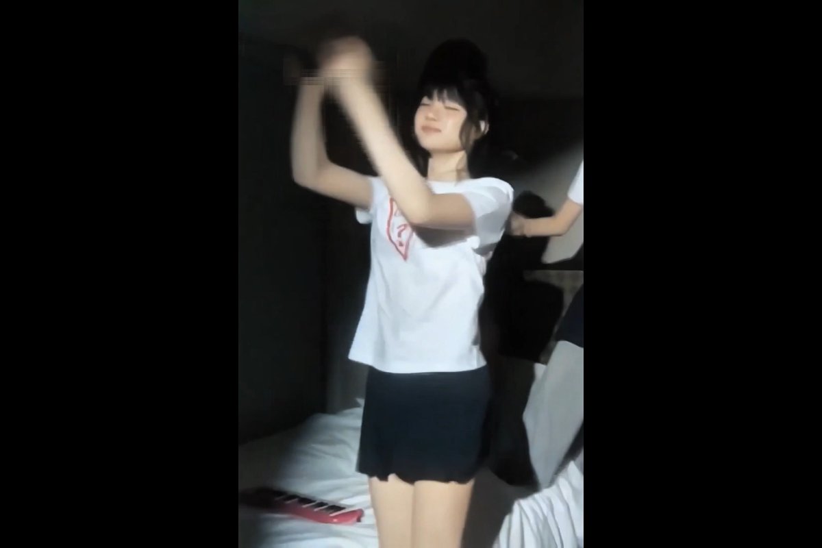 A blurry video still captures a woman in a white t-shirt with a red logo and a black mini skirt, standing in a dimly lit room with her arms raised near her head.