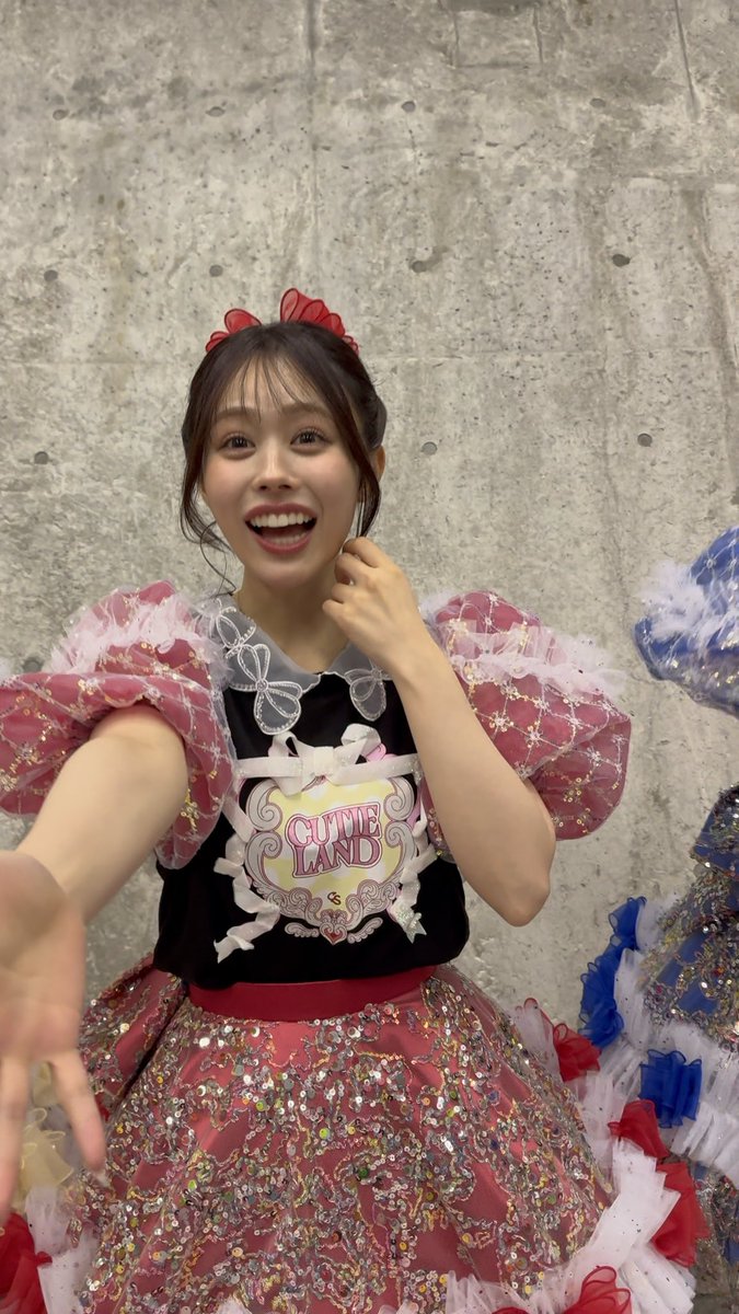 A young woman beams at the camera while wearing a detailed pink, red, and black idol costume featuring the text 'CUTIE LAND'.