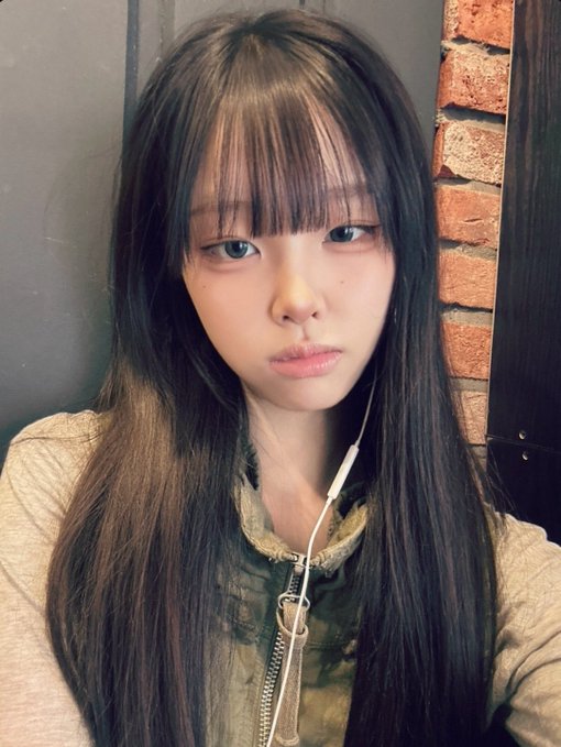 A selfie of a young woman with long straight black hair and bangs, wearing white earphones and a beige jacket over a green top, posed against a brick wall.