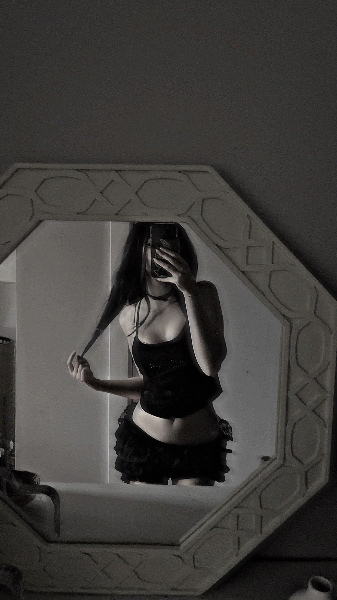 A black-and-white vertical shot showing a mirror reflection of a person in a black ruffled outfit holding a strand of hair, framed by an octagonal mirror.
