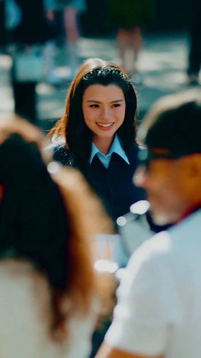 A smiling woman with dark hair wearing a dark blazer and light blue shirt stands among a blurred crowd in an outdoor setting.
