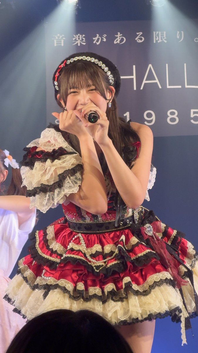 A young Japanese idol performs on stage in an elaborate red, white, and gold ruffled dress, singing into a microphone with Japanese text visible on the backdrop.