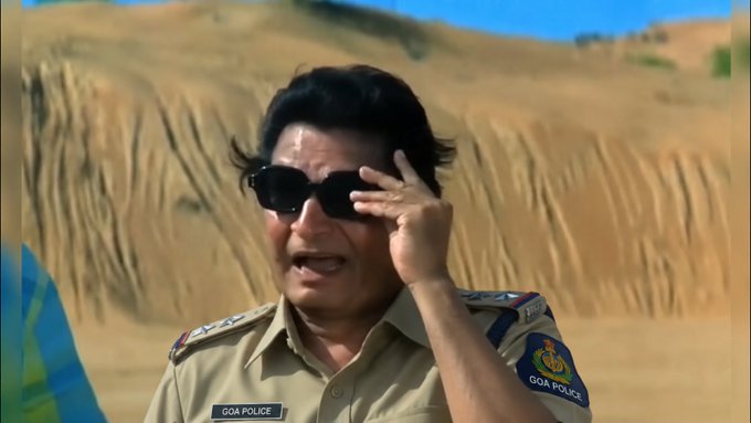 A screenshot from a video showing a Goa Police officer in uniform wearing sunglasses, gesturing with his hand while standing in front of a sandy embankment.