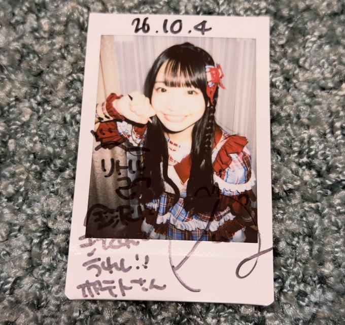 A signed polaroid photograph dated '16.10.4' showing a smiling young woman with long dark hair wearing a red and plaid outfit with a red bow, featuring handwritten Japanese signatures and messages in black ink, resting on a gray carpet.