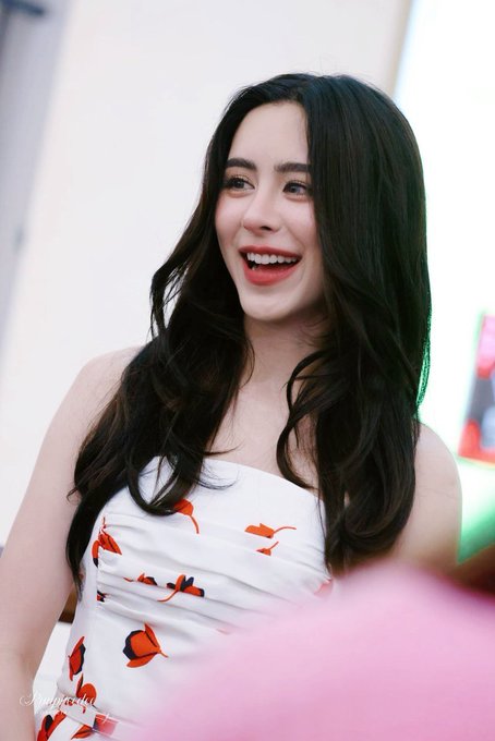 A portrait of a young woman with long dark hair wearing a white dress with orange floral patterns, smiling brightly with red lipstick.