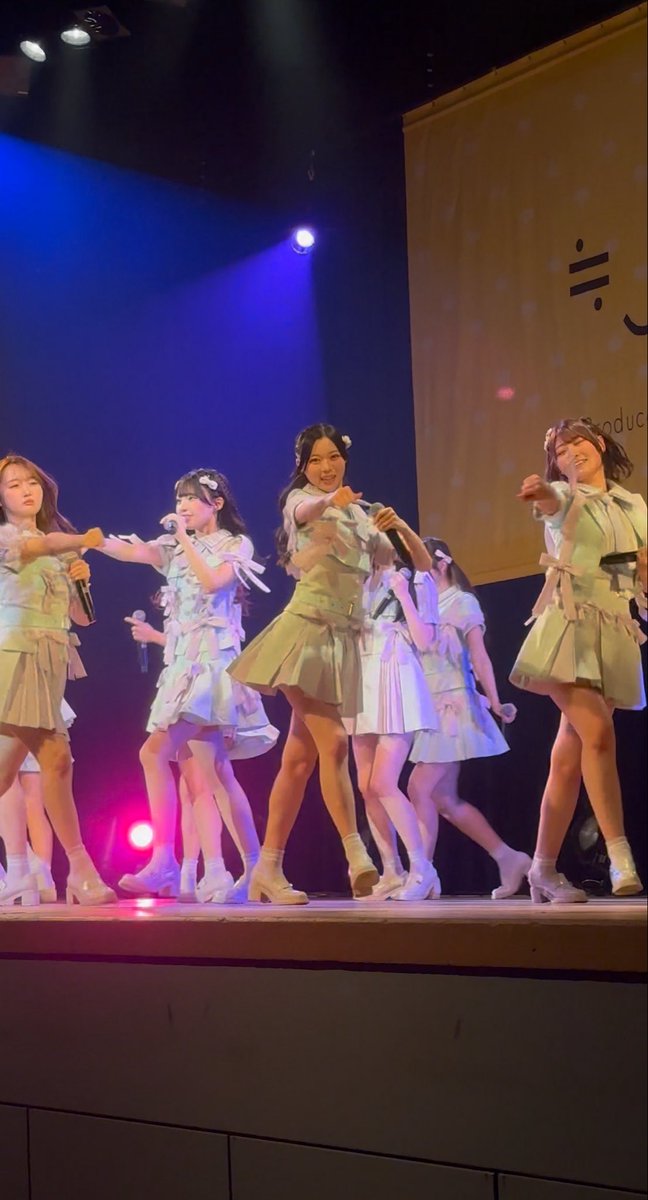 A group of young female performers in matching white and pastel-colored outfits dance on a stage illuminated with blue lighting.