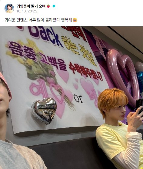 A screenshot of a Korean social media post (귀염둥이 뭉가 오빠) dated 10.18, 20:25, with text '귀여운 컨텐츠 너무 많이 올라왔다 행복해' (Happy that so much cute content has been posted) above a photo of a person with orange hair standing next to a decorated banner with hearts.