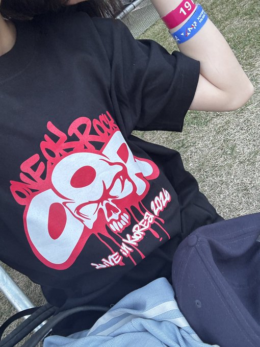 A close-up of a black "ONE OK ROCK" t-shirt with a red skull graphic and "LIVE IN KOREA 2024" text, worn by someone with festival wristbands ("19", "AIS").
