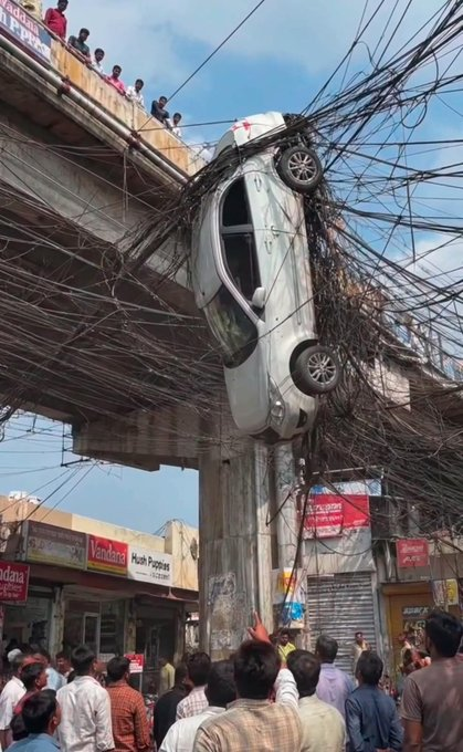 Shocking photograph of a white car hanging off the edge of a concrete bridge/entrance, entangled in a massive tangle of electrical wires, with a crowd of onlookers gathered below.