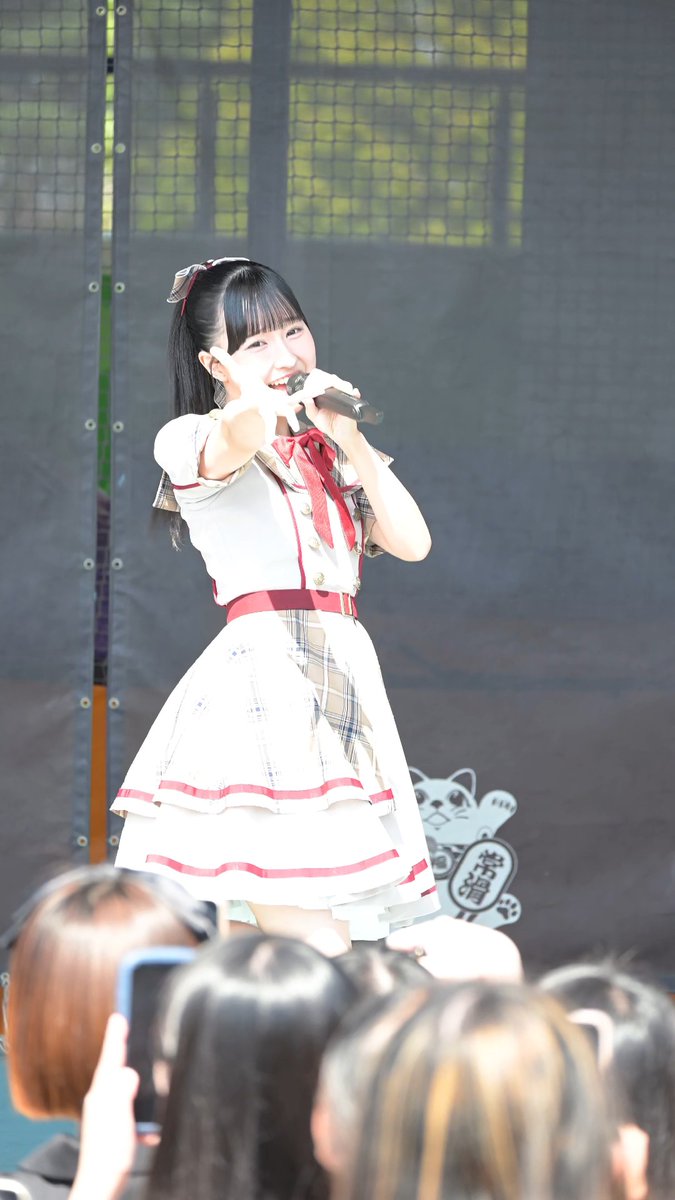A young female idol with twin tails, wearing a white and red plaid dress, performs with a microphone on an outdoor stage in front of a crowd.