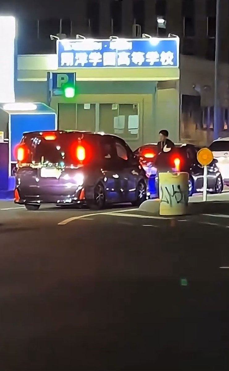 A frustrated driver yells at a honking motorist outside a Japanese driving school (明洋学園高等学校) at night, while standing next to a black car near a parking sign.