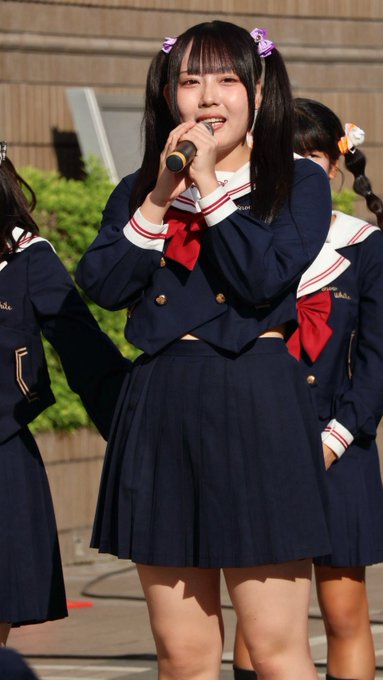 A smiling Japanese idol wearing a navy blue sailor-style uniform with red bows and hair ribbons sings into a microphone during an outdoor performance.