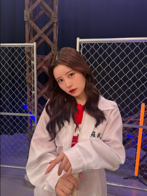 A young woman with long dark hair posing in a white jacket with red accents and Japanese text, standing in front of a chain-link fence and scaffolding.