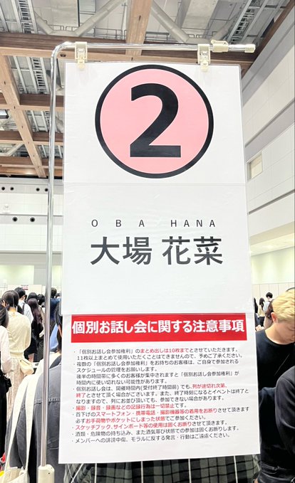 A sign for 'OBAHANA 大場花菜' (member #2) displays '個別お話し会に関する注意事項' (Notices regarding individual talk events) with bullet points of rules for an idol handshake/talk event.