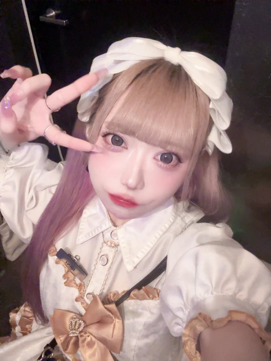 A selfie of a person in cosplay with light brown and pink hair, a large white bow headband, and a white outfit with gold accents, making a peace sign near their face.