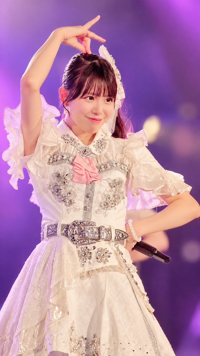 A young woman in an elaborate white dress with ruffles and a jeweled belt poses on stage with her hand above her head.