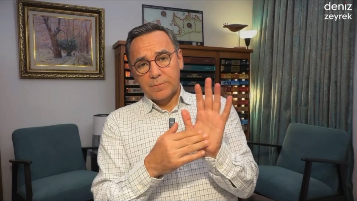 A man wearing glasses and a checkered shirt sits in a room with bookshelves and a painting, gesturing with his hands; the watermark 'deniz zeyrek' appears in the corner.