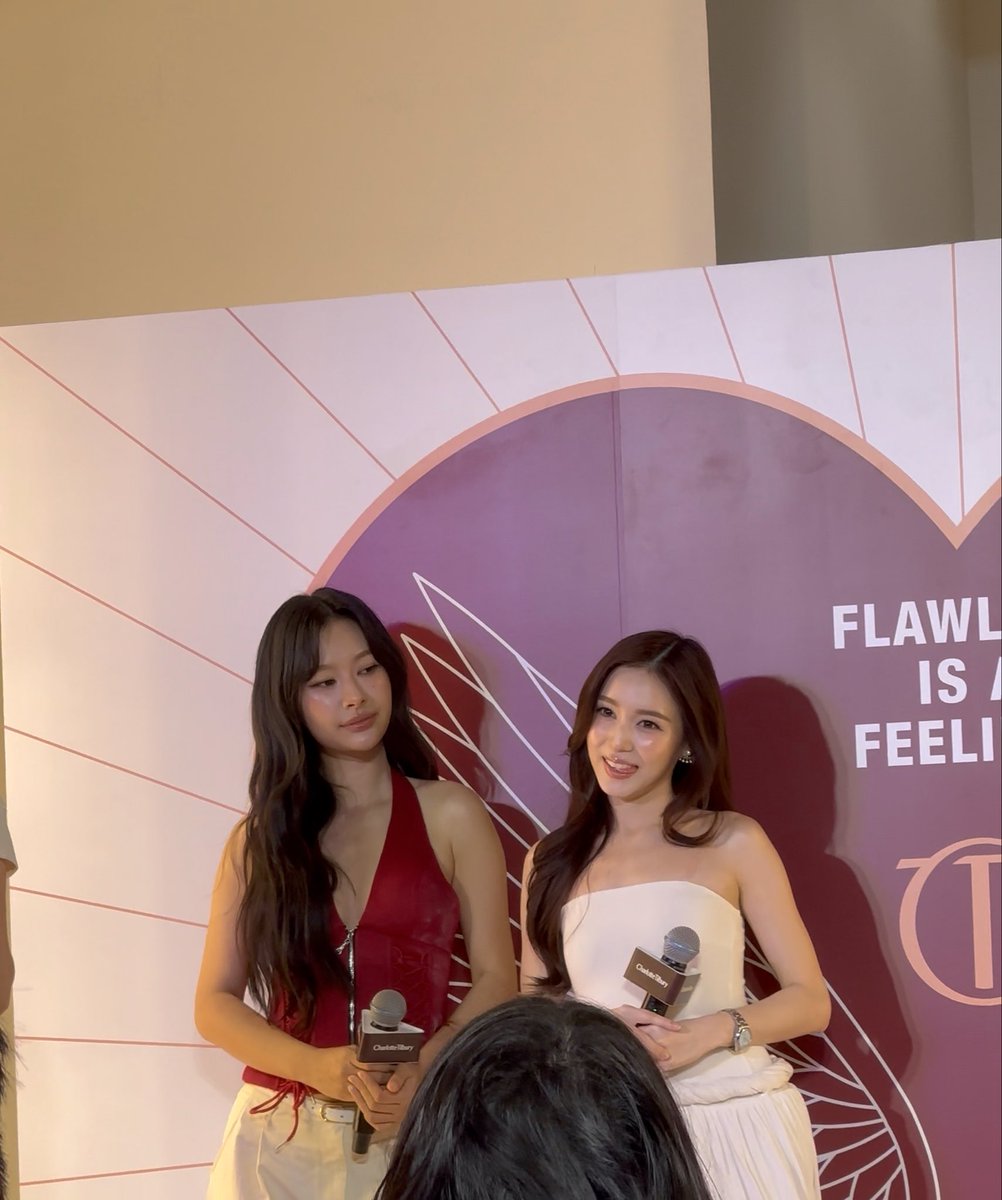 Two women holding microphones at a Charlotte Tilbury event, one in a red halter top and the other in a white strapless dress, standing before a backdrop reading 'FLAWLESS IS A FEELING'.
