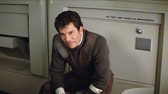 A man in a dark uniform crouches down, looking directly at the camera, next to a wall fixture labeled "DO NOT USE WHILE IN SPACEDOCK"—the only toilet shown in any Star Trek film, appearing in Star Trek V: The Final Frontier.