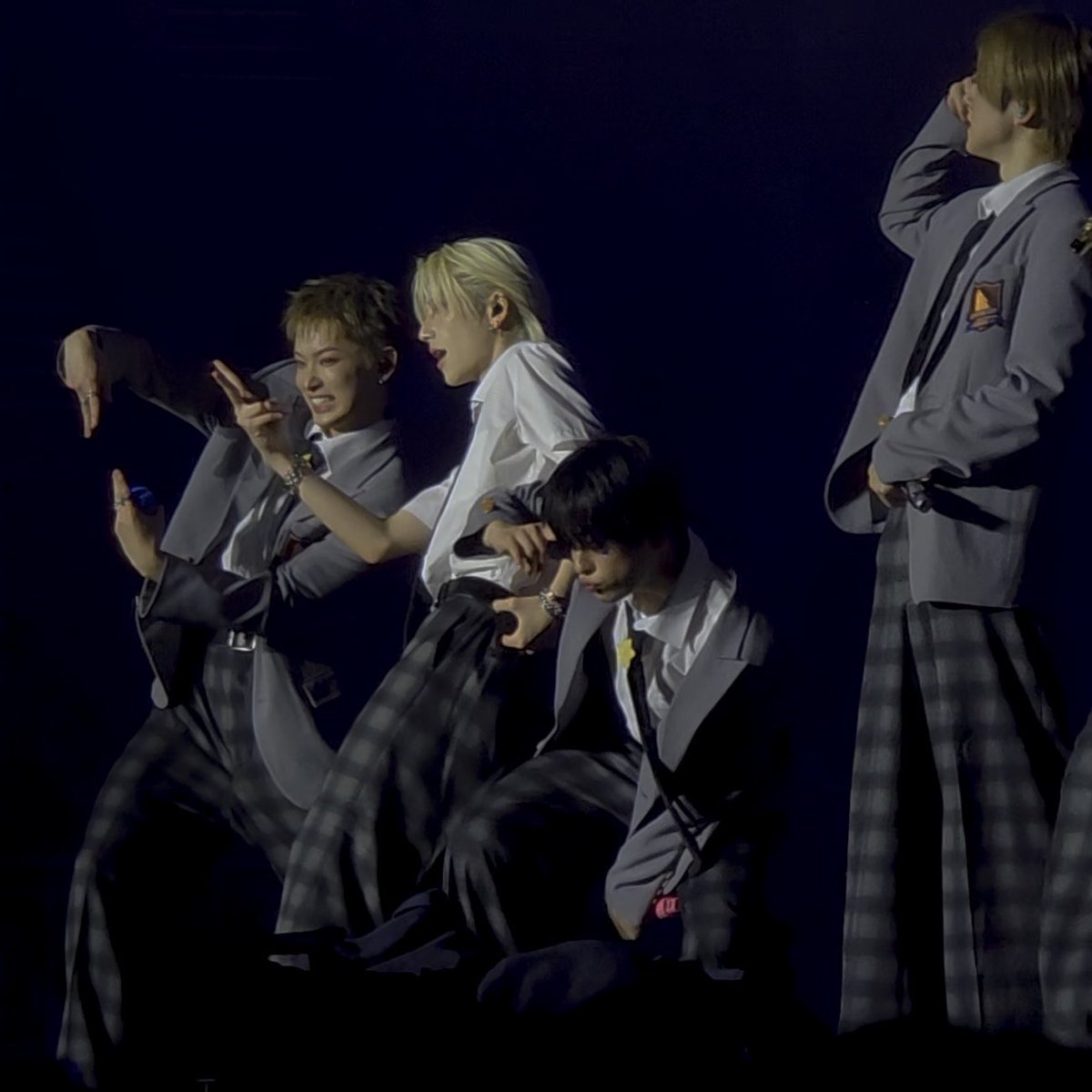 Four young male performers in matching gray blazers, white shirts, ties, and plaid pants pose dynamically under a spotlight on a dark stage.