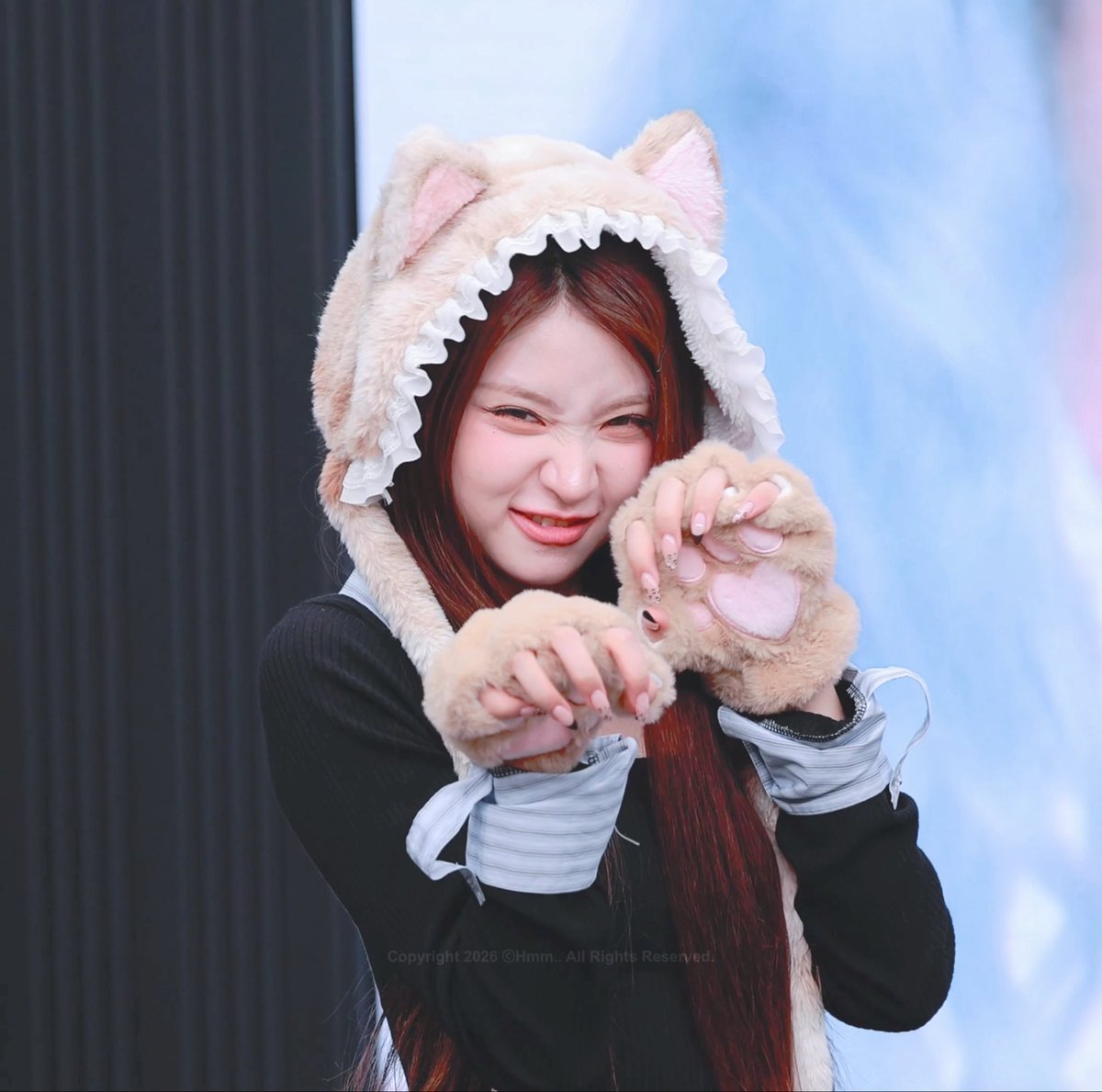 A smiling woman with long red hair wears a furry beige cat-ear hood with paw gloves, holding her hands up playfully for a fan photo.