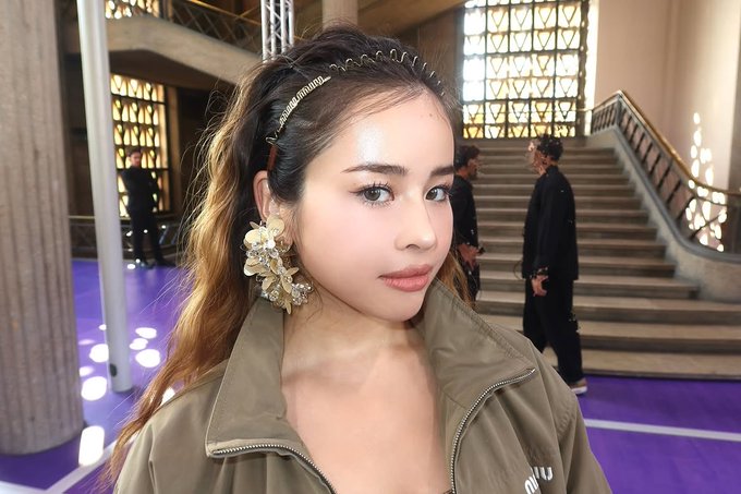 A close-up portrait of a young woman with long brown hair, wearing a decorative headband, large flower earrings, and an olive green jacket, photographed at an indoor event with a purple floor.