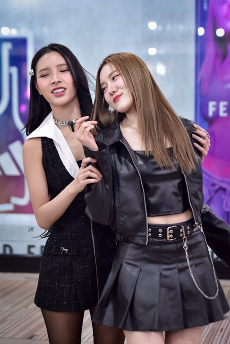 Two young women posing together at what appears to be a K-pop event, one in a black vest outfit with a bow hair clip and the other in a black leather jacket and pleated skirt with a chain belt.