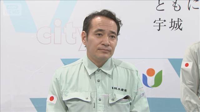 A man in a light green work uniform with a Japanese flag patch sits in front of a backdrop displaying Japanese text including '宇城' and the letters 'city'.