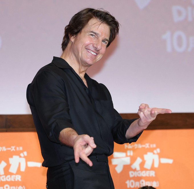 Tom Cruise smiles broadly and flashes a gal peace sign while promoting his film "DIGGER・Digger" at a press conference in Tokyo, with Japanese promotional banners visible in the background indicating the film's October 9 release date.