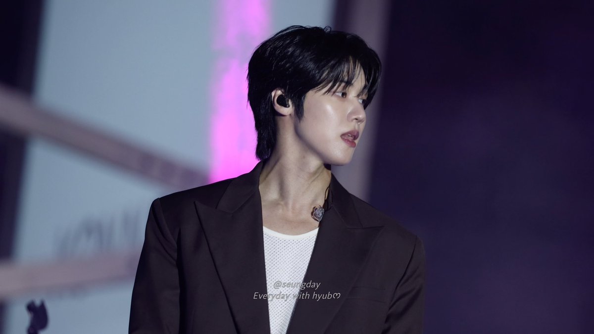 A young man with black hair wearing a dark blazer over a white mesh top and an in-ear monitor, performing on stage; watermark reads "@seungday Everyday with hyub♥".