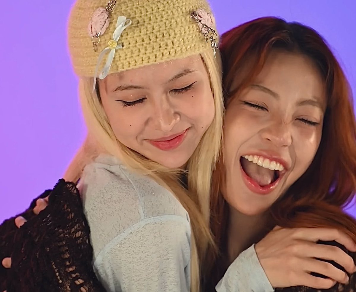 Two young women pose closely together against a purple background; the blonde on the left wears a cream beanie with charms and smiles softly with closed eyes, while her friend on the right laughs joyfully with her mouth wide open.