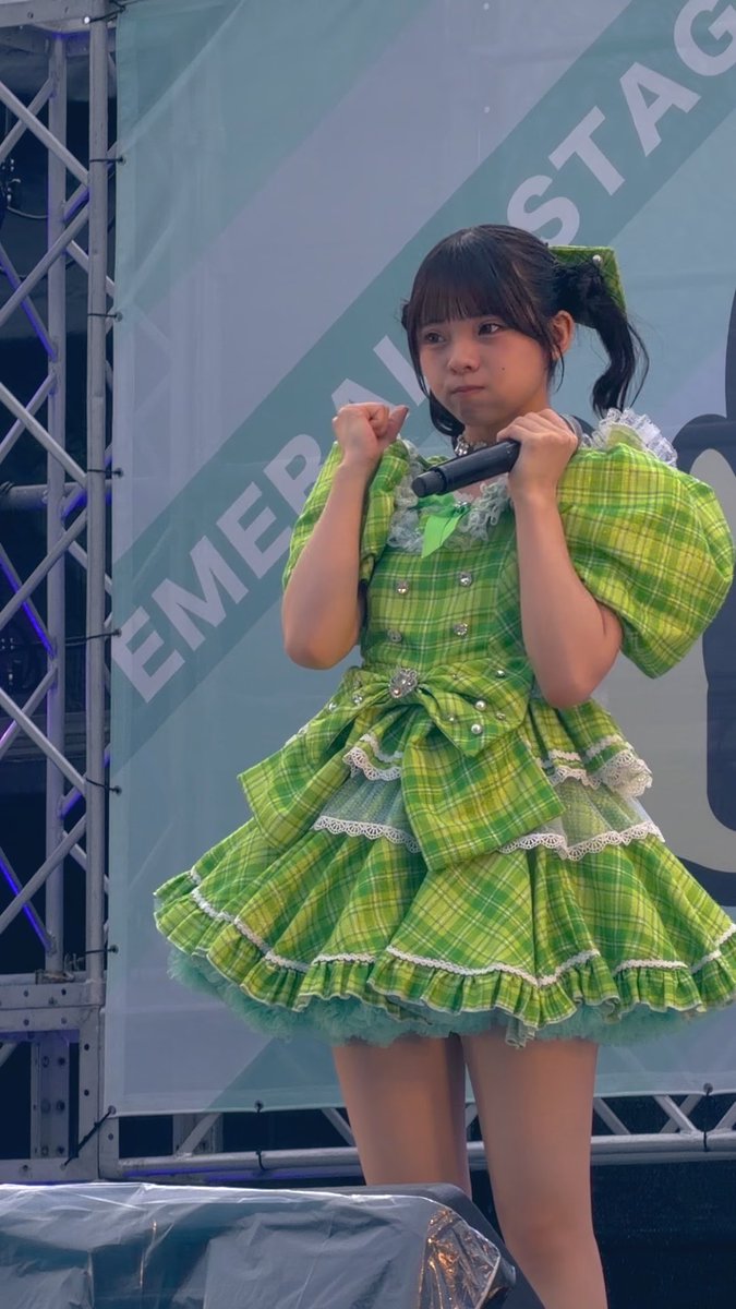 A young woman in a vibrant green plaid ruffled dress stands on stage holding a microphone, with a banner reading 'EMERGING STAGE' visible behind her.