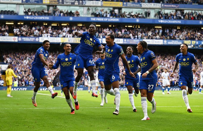 Chelsea FC players in blue jerseys with 'USDC' sponsorship logos celebrate on the field at Stamford Bridge, with 'THE PRIDE OF LONDON' and 'WORLD CHAMPIONS' banners visible in the background.